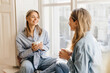 © Look! - Two pleasant young caucasian women are talking drinking cappuccino sitting on windowsill in morning. Blondes wear casual clothes. Relax, leisure at home, indoors.
