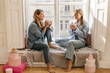 © Look! - Two joyful european ladies in good mood drink coffee and laugh sitting on windowsill warm day. Blondes wear casual clothes barefoot. Lifestyle concept