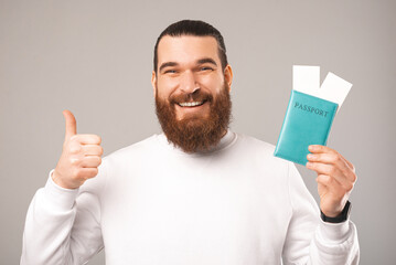 Wall Mural - Close up photo of a bearded man showing thumb up while holding a passport. Studio shot over grey background.