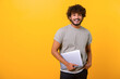 © Vadim Pastuh - Cheerful Indian young man holding laptop standing isolated on yellow, looking at camera with happy smile. Male student or freelancer carrying laptop computer. Copy space