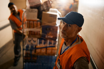 Poster - Mature warehouse worker and his coworker moving rack with packages through storage hallway.