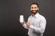 © Vadim Pastuh - Mock-up concept. Handsome confident bearded Indian man in formal wear showing smartphone with empty screen, points finger at blank device display isolated on black, male advertising new mobile app