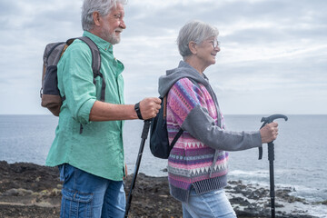 Active smiling caucasian senior couple hiking with help of stick on the cliffs, elderly white haired people enjoying sea excursion and healthy lifestyle. Horizon over water