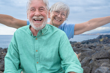 Happy senior caucasian couple while sitting outdoors at sea laughing at the camera, woman with outstretched arms. Two seniors enjoying the freedom and relaxing in retirement. Horizon over the water