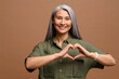 © Vadim Pastuh - Love you. Portrait of attractive romantic mature woman standing and making heart with hands, while smiling playfully. Indoor studio shot isolated on brown background