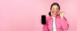 Portrait of asian girl showing mobile phone screen, reacting surprised, standing over pink background