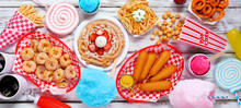 Carnival Theme Food Table Scene Over A White Wood Banner Background. Top View. Summer Fair Concept. Corn Dogs, Funnel Cake, Cotton Candy And Snacks.