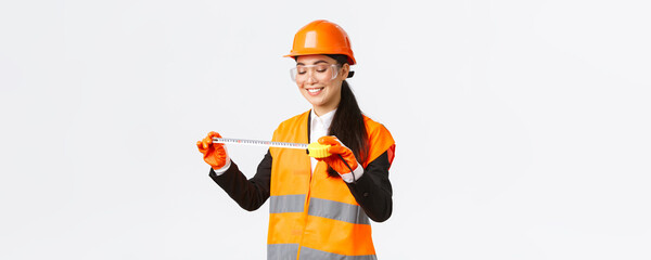 Wall Mural - Pleased smiling asian female architect achieve good results, looking satisfied at tape measure after measuring layout at construction area, standing in safety helmet and reflective clothing