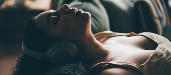 Wall Mural - Curly haired overweight young woman wearing comfortable clothes lies on green sofa listening to music in wireless headphones in stylish room