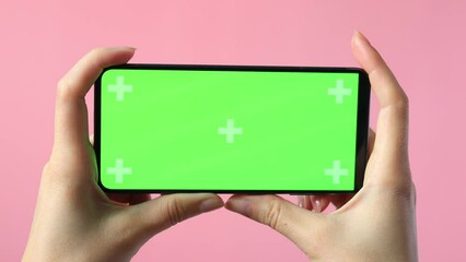 close-up of female hands holding smartphone with blank green screen showing copy space for app or website design, on pink background , mockup.