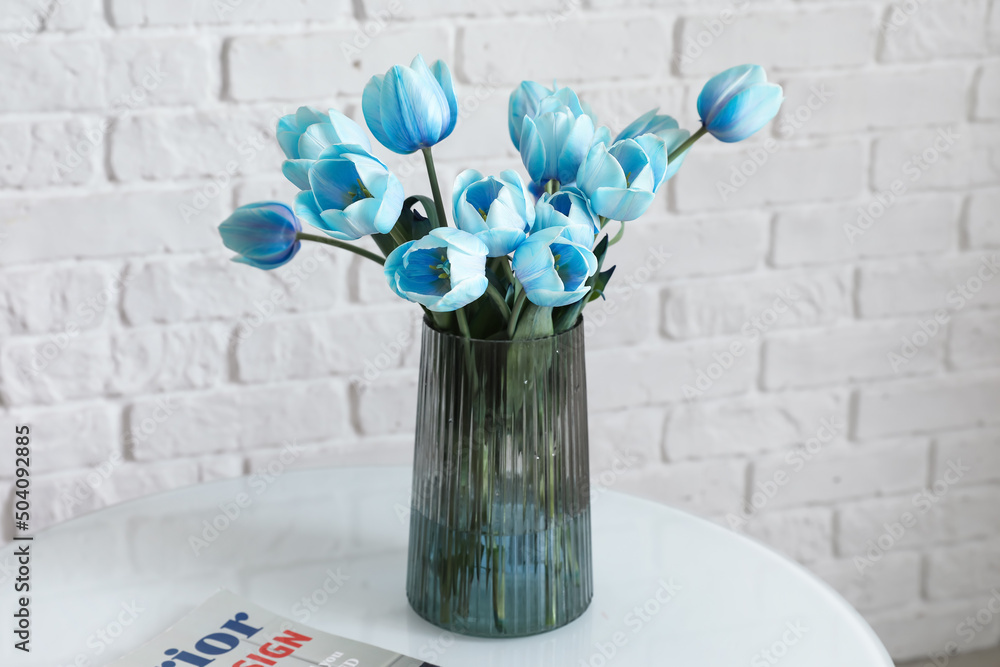 Vase with blue tulips and magazine on table near white brick wall