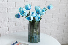 Vase With Blue Tulips And Magazine On Table Near White Brick Wall