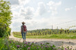 Woman with brown hair, gray t-shirt, jeans, red backpack and straw hat hiking in the nature, beautiful flowers and a wine field next to her, Auerbach, Bensheim, Germany