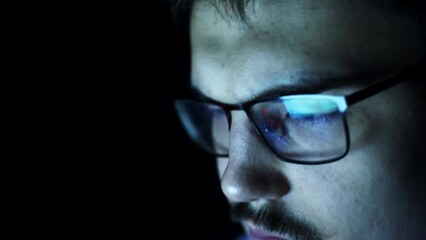 Very tired nerdy student with glasses is sitting at darkness browsing internet, closeup face. Closeup reflection in glasses of man browsing website on computer in dark