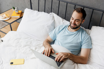 Wall Mural - Top view young happy man wear blue t-shirt lying in bed hold use work on laptop pc computer rest relax spend time in bedroom lounge home in own room house wake up dream. Good mood bedtime concept