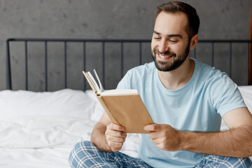 Young smiling happy man wear casual blue t-shirt sitting on bed read novel book learn new information rest relax spend time in bedroom lounge home in own room house wake up dream. Real estate concept.