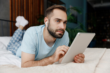 Wall Mural - Full body pensive young man in blue t-shirt lying on bed listen music in earphones use tablet pc computer rest relax spend time in bedroom lounge home in own room house dream. Morning mood concept