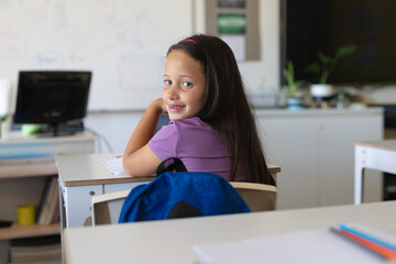 Portrait of caucasian elementary schoolgirl looking over shoulder while sitting at desk in class