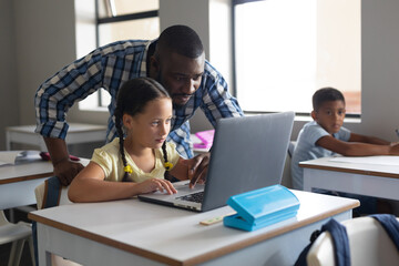 Wall Mural - African american young male teacher showing laptop to caucasian elementary schoolgirl at desk