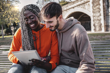 Multi-ethnic couple of young people using digital tablet device browsing content sitting outdoors in the park on a bench - lifestyle concept of biracial couple having fun using tech