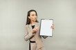 © bodnarphoto - Business woman with a tablet in her hands stands on a beige background looks at a blank white sheet of paper and shows a pen on copy space