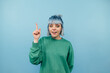 Positive lady with blue hair stands on a blue background with a happy face looking at the camera and showing finger up