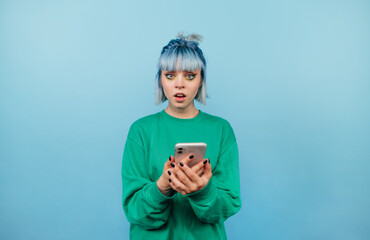 Wall Mural - Surprised hipster girl with blue hair stands on a blue background with a smartphone in his hands and looks at the camera with a shocked face.