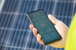 Unrecognizable person showing a solar power generation smartphone app with solar panels in the background.