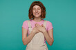 Indoor portrait of young ginger female, wears overalls and pink t shirt posing over blue background keeps her eyes closed and hands on heart, smiles broadly