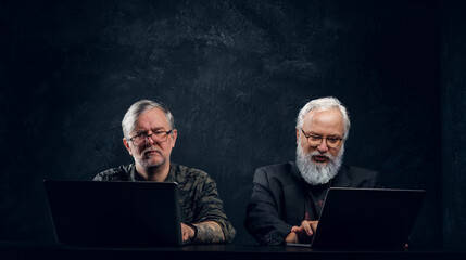 Portrait of two senior men coworkers using laptops sitting at table against dark background.