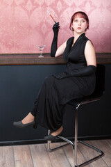 leggy woman wearing long black dress and long black gloves is sitting on a bar chair with glass of c