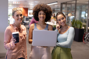 Wall Mural - Portrait of happy biracial businesswomen standing with laptop and drink at modern workplace