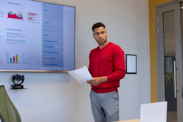 Wall Mural - Hispanic male advisor holding document looking away while standing by business plan at workplace