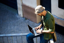 From Above White Man In Casual Clothes And Hat Sitting On Balcony Railing And Using Laptop For Work On Sunny Day In City.