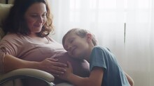 Child Hugs And Kisses Her Mother's Pregnant Belly. Happy Son Strokes The Big Belly His Pregnant Mom Near Window At Home. Happy Family Life In An Expectation A Newborn. Pregnancy And Maternity.