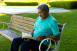 © wavebreak3 - Focused biracial senior man wearing blue t-shirt reading book while sitting on bench in park