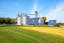 Solar Panels And A Grain Elevator With Big Steel Storage Bins In A Modern Farm On A Sunny Autumn Day. Use Of Renewable Energy In Agriculture Concept.