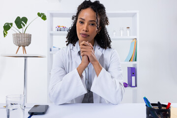 Wall Mural - Portrait of confident african american mid adult female doctor with hands clasped sitting at desk