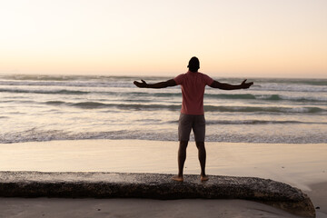 Rear view of african american young man with arms outstretched standing at beach against clear sky