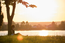 Lonely Woman Sitting Alone On Green Grass Lawn Leaning To Tree Trunk On Lake Shore On Warm Evening. Solitude And Relaxing In Nature Concept