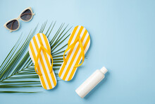 Summer Vacation Concept. Top View Photo Of Yellow Striped Flip-flops Sunglasses Sunscreen Bottle And Palm Leaves On Isolated Pastel Blue Background