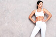 © halayalex - Fitness confident woman in white sports clothing. Sexy young beautiful model with perfect body. Female posing near grey wall in studio. Stretching out before training