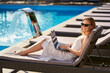 © artiemedvedev - Self employed woman telecommuting with team on project from tropical island resort. Freelancer businesswoman doing remote work with laptop on swimming pool beach bed at spa. Girl tanning and relaxing.