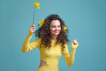 Happy Curly Brunette Girl In A Yellow Dress With Daffodil Flowers In Her Hair Isolated On A Blue Background.