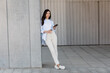 © LIGHTFIELD STUDIOS - full length of brunette woman standing with smartphone and coffee to go near grey wall.