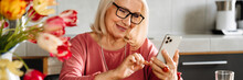 A Positive Elderly Woman In Glasses Leafing Something On The Phone In The Kitchen