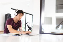 Asian Teenage Boy Wearing Headphones With Microphone On Table Using Laptop While Podcasting At Home