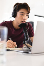 Asian Teenage Boy Writing In Diary While Podcasting Over Microphone And Laptop On Table At Home