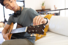 Asian Teenage Boy Adjusting Guitar Strings While Sitting On Sofa In Living Room At Home, Copy Space