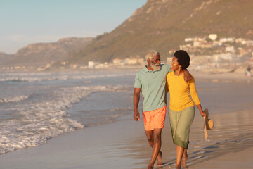 African american senior man with hand on mature woman's shoulder walking at beach at sunset